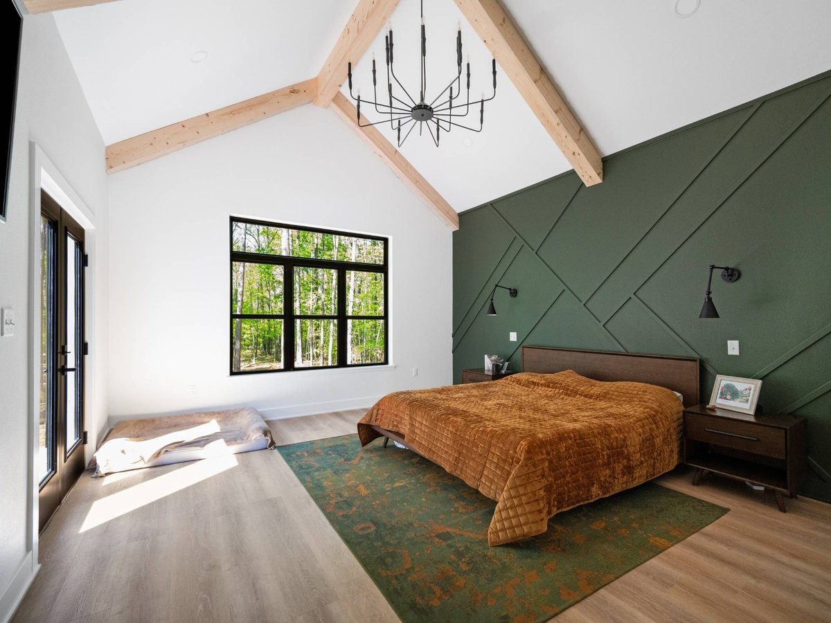 Photo of a primary bedroom with vaulted wood beams, green accent wall, and large black-framed windows in East Texas.