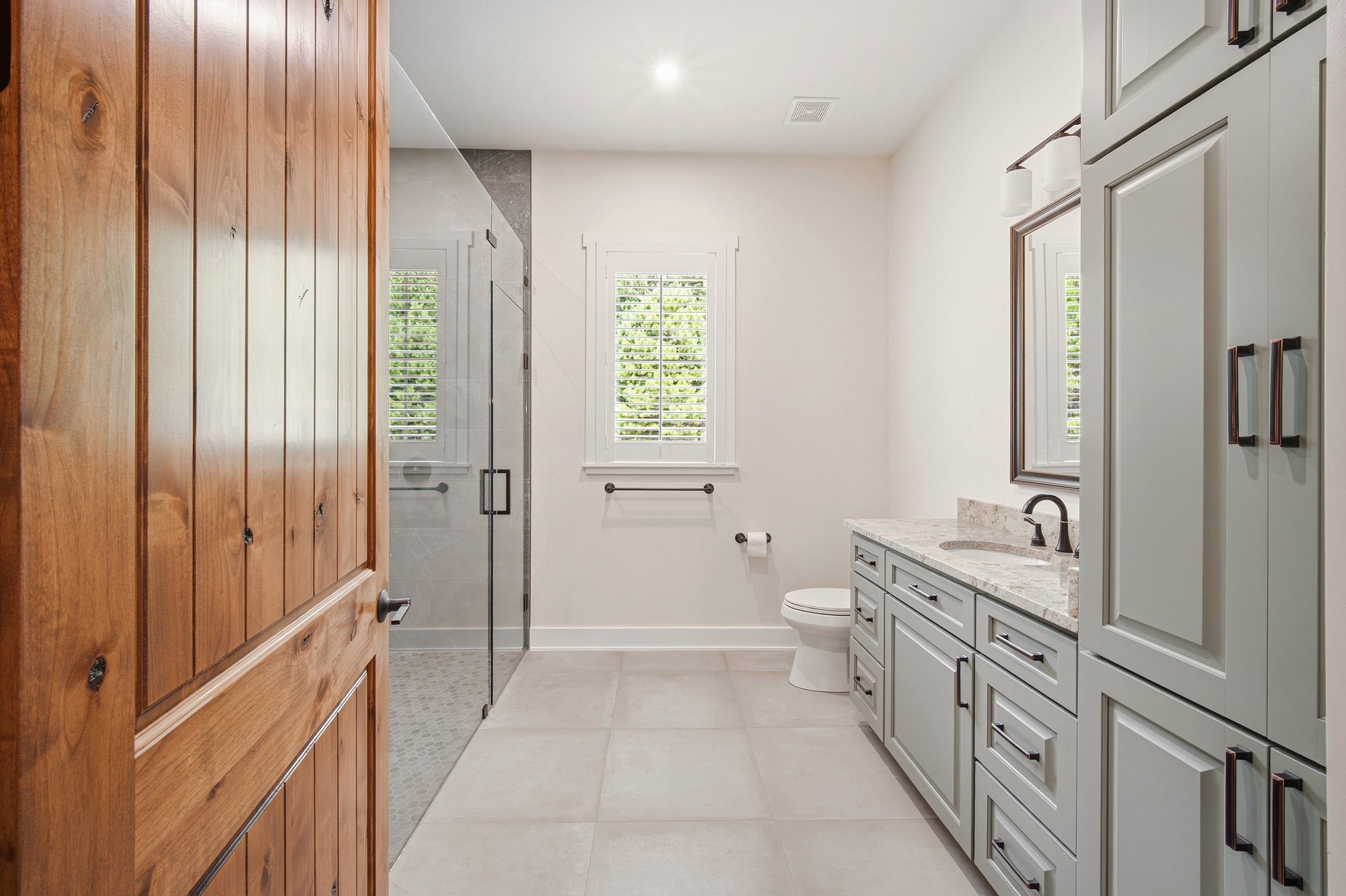 Bathroom in Bort Custom Homes’ Henderson, TX residence, with a stained wood door, gray vanity cabinets, and a glass-enclosed shower.