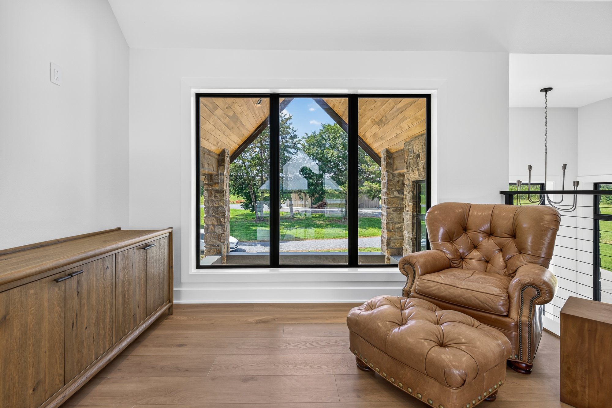 Upstairs sitting nook with a tufted leather armchair and ottoman, wood credenza, and a large window looking out to a stone-and-timber covered porch.